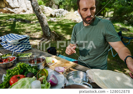 Man Preparing Outdoor Picnic Meal with Fresh Ingredients in a Forest Setting 128012878