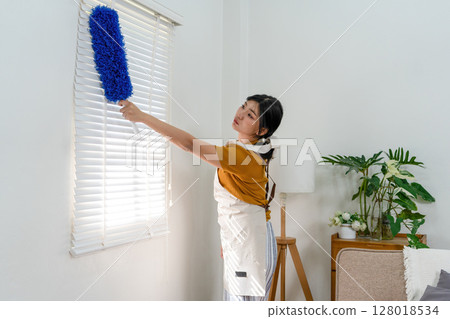 Young modern Asian woman doing house cleaning at home. Confident and independent homemaker maintaining a clean, stylish living space. Young modern Asian woman doing house cleaning at home. Confident and independent homemaker maintaining a clean, stylish living space. 128018534