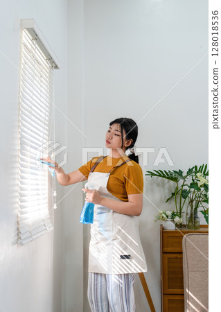 Young modern Asian woman doing house cleaning at home. Confident and independent homemaker maintaining a clean, stylish living space. Young modern Asian woman doing house cleaning at home. Confident and independent homemaker maintaining a clean, stylish living space. 128018536