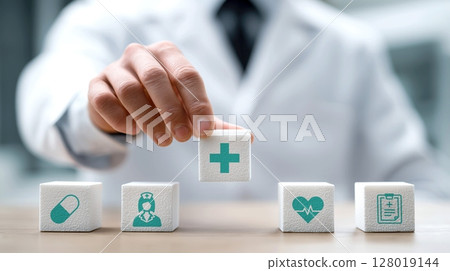 Doctor arranges white blocks with medical symbols, including cross, pill, nurse, heart, and clipboard, symbolizing healthcare concepts 128019144