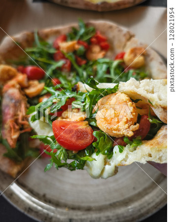 Hand holding Gourmet pizza featuring succulent shrimp, peppery arugula, and ripe cherry tomatoes on rustic surface in aosta, italy 128021954