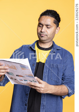 Indian young guy browsing though financial audit report paperwork, feeling overworked and confused of the balance numbers. Accountant person standing against studio background. Indian young guy browsing though financial audit report paperwork, feeling overworked and confused of the balance numbers. Accountant person standing against studio background. 128026276