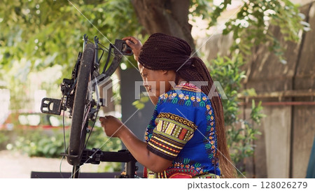Determined female cyclist using specialized work tool to repair and adjust bike rear derailleur. Sporty african american woman mending her bicycle with professional multitool in home yard. 128026279
