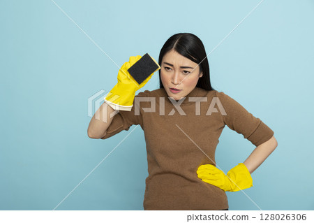 Portrait of serious asian woman in yellow rubber gloves, holding cleaning sponge and hand on waist, staring ahead with intent. Housemaid standing ready for chores with confidence and precision. Portrait of serious asian woman in yellow rubber gloves, holding cleaning sponge and hand on waist, staring ahead with intent. Housemaid standing ready for chores with confidence and precision. 128026306