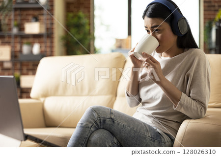 Female entrepreneur wearing headphones and sipping tea while watching online webinar on laptop. Remote employee with wireless headset and coffee cup in hand, enjoying break on living room sofa. 128026310