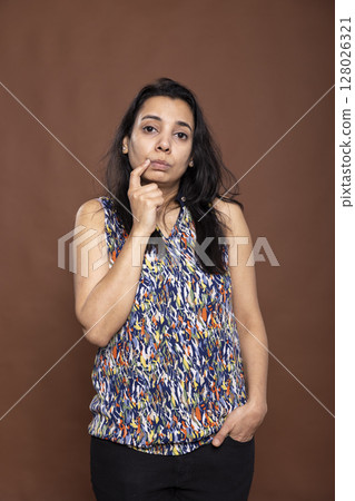 Contemplative woman standing with thoughtful expression, touching her chin and pondering an answer. Female in casual outfit appearing curious and doubtful, isolated in brown studio background. 128026321