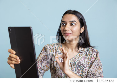 Confident upbeat girl sharing a story with best friend over web video call, standing against blue background with smartphone. Happy woman talking with mate online on teleconference. Confident upbeat girl sharing a story with best friend over web video call, standing against blue background with smartphone. Happy woman talking with mate online on teleconference. 128026328