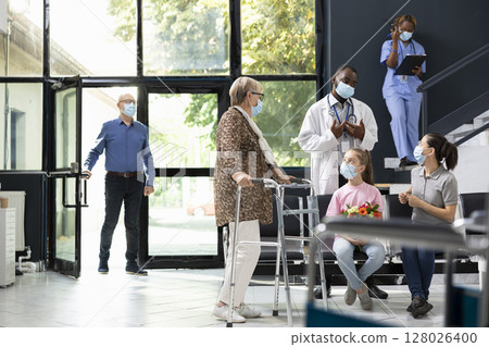 Girl visiting senior grandmother using walking frame at hospital lobby, offering flowers with love and care while respecting medical appointment safety regulations and health assurance policies. Girl visiting senior grandmother using walking frame at hospital lobby, offering flowers with love and care while respecting medical appointment safety regulations and health assurance policies. 128026400