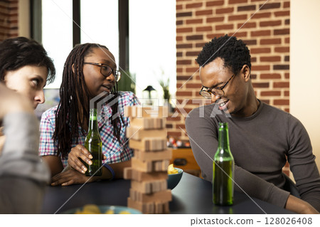 African american couple drinking beer and making goofy facial expressions in brick wall room. Group of friends at table, enjoying board games and relaxed bonding time with wooden game pieces. African american couple drinking beer and making goofy facial expressions in brick wall room. Group of friends at table, enjoying board games and relaxed bonding time with wooden game pieces. 128026408