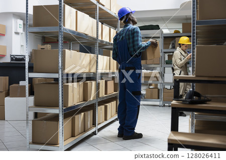 Black male employee examine labeled parcels on shelves in a storage unit depot. Logistics services support bulk shipping and retail inventory movement for supply chain, storehouse. Black male employee examine labeled parcels on shelves in a storage unit depot. Logistics services support bulk shipping and retail inventory movement for supply chain, storehouse. 128026411