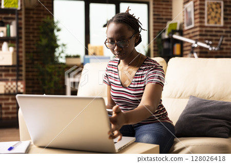 Focused woman works from home, reviewing company growth analytics on laptop. African American female entrepreneur touching device screen and reading online data with full concentration. 128026418