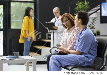 Medical assistant showing glucose levels test measurement for diabetes routine checkup in hospital hallway. Asian woman receives usage tips on glucometer, blood sugar readings. 128026463
