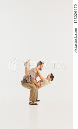Retro couple performing lively swing dance move in vintage-inspired attire against white background 128026470