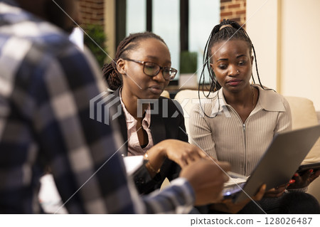 African American woman wearing glasses, using laptop to give business project updates to her colleagues. Female coworkers seated in office couch, discussing marketing strategies and solutions. 128026487