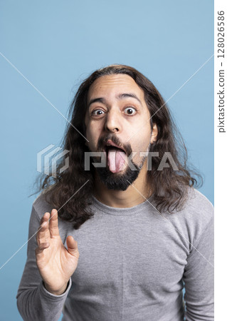 Goofy young adult with long hair and beard poses playfully in the studio, expressing positive emotions and carefree humor. Joyful person showcases unique, comical personality and fun spirit. Goofy young adult with long hair and beard poses playfully in the studio, expressing positive emotions and carefree humor. Joyful person showcases unique, comical personality and fun spirit. 128026586