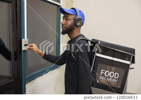 African american worker approaches the doorway to drop off a hot meal order, ringing the intercom to deliver the food. Young adult employee ensuring an express delivery takeaway service. African american worker approaches the doorway to drop off a hot meal order, ringing the intercom to deliver the food. Young adult employee ensuring an express delivery takeaway service. 128026631