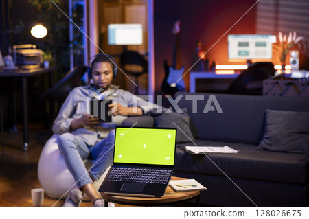 African american guy listening to music on headphones next to green screen, feeling relaxed sitting on the bean bag in the living room. Happy chill man sitting in his trendy loft. African american guy listening to music on headphones next to green screen, feeling relaxed sitting on the bean bag in the living room. Happy chill man sitting in his trendy loft. 128026675