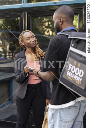 Black woman paying for her takeaway meal order with a contactless card at the doorstep. Delivery man carrying a thermal bag, completes fast food transaction using a wireless POS terminal. 128026700
