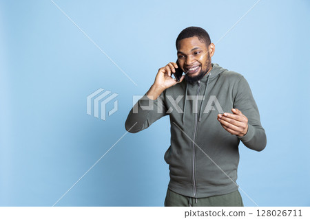 African american young adult enjoys talking to his friends on phone call using smartphone, poses against studio background. Relaxed guy catching up with mates during telephone call. African american young adult enjoys talking to his friends on phone call using smartphone, poses against studio background. Relaxed guy catching up with mates during telephone call. 128026711