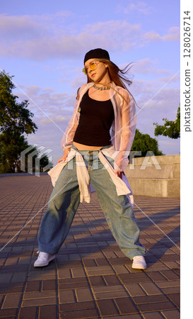 Young woman dancing energetically outside in casual urban wear at sunset 128026714