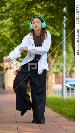 Inner rhythm. Young woman dancing with eyes closed, on tree-lined street wearing headphones 128026772