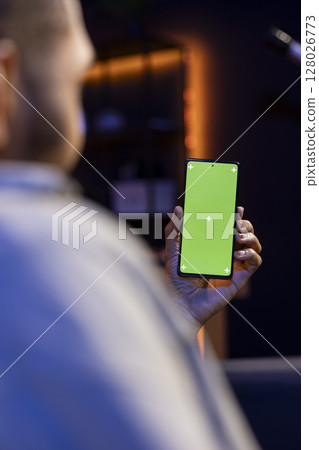 Black remote worker looking at green screen on his device at home, relaxing in the living room and solving his freelancing tasks. Young adult being casual in his trendy apartment, teleworking. 128026773