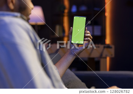 African american remote worker uses chroma key screen on gadget to work in the living room, sitting on the couch in his modern stylish living room. Happy young adult on the internet. African american remote worker uses chroma key screen on gadget to work in the living room, sitting on the couch in his modern stylish living room. Happy young adult on the internet. 128026782