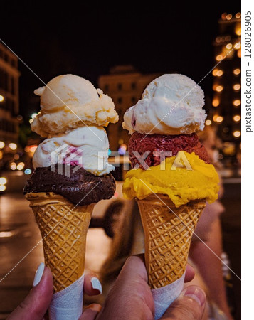 Ice cream in a cone of different flavors and colors, on the street in the evening in Rome, held in the hands of young people, vertical photo 128026905