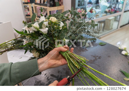 Close-up of florist trimming flower stems with pruning shears while holding a wrapped bouquet of white and peach flowers with eucalyptus greenery Close-up of florist trimming flower stems with pruning shears while holding a wrapped bouquet of white and peach flowers with eucalyptus greenery 128027674