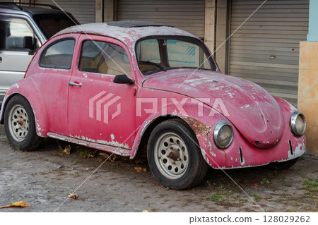 Front view of an old, rusty pink Volkswagen Beetle parked outside 128029262