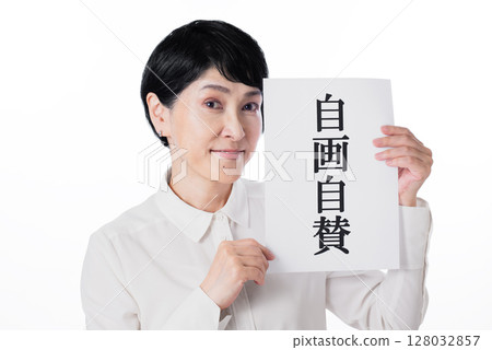 A middle-aged woman with short hair wearing a white shirt and smiling while holding a piece of paper with the words "Self-praise" written on it 128032857