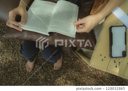 Woman enjoying digital detox weekend retreat while reading book on outdoor hotel seating with bare feet on stone floor for wellness and mental health self care relaxation therapy. Disconnection. 128032865