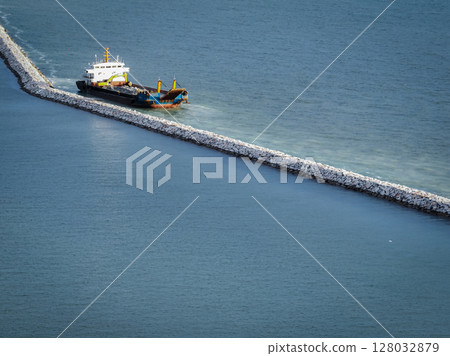 Marine construction vessel working on stone breakwater project in ocean. Coastal infrastructure development and harbor protection engineering work in progress. Port construction and coastal defense. 128032879