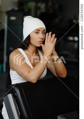 Portrait confident beautiful young brunette woman is wearing white suit and cap sitting in the gym 128034560