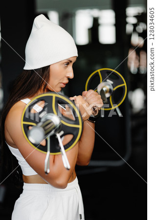 A young woman does exercises for biceps with a barbell with a curved bar, standing in the gym. The woman in the gym is wearing white suit is doing workout A young woman does exercises for biceps with a barbell with a curved bar, standing in the gym. The woman in the gym is wearing white suit is doing workout 128034605