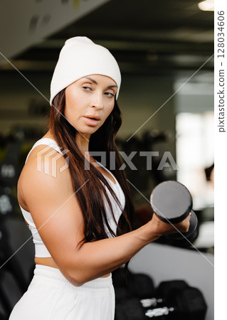 Young fit woman practicing dumbbell press while standing in the gym. Slim female in sportswear exercises with weights in a gym Young fit woman practicing dumbbell press while standing in the gym. Slim female in sportswear exercises with weights in a gym 128034606