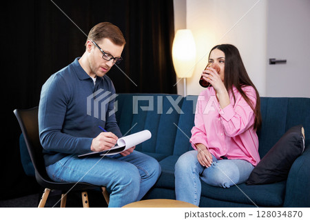A young psychologist conducts a psychotherapy session for a woman who has psychological problems. A woman sits on a sofa and holds a glass of water in her hands 128034870