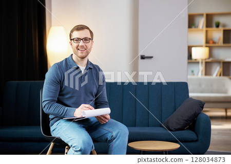 Portrait of happy male psychologist looking at camera and taking notes during therapy session at clinic. Friendly psychotherapist posing and smiling at office 128034873