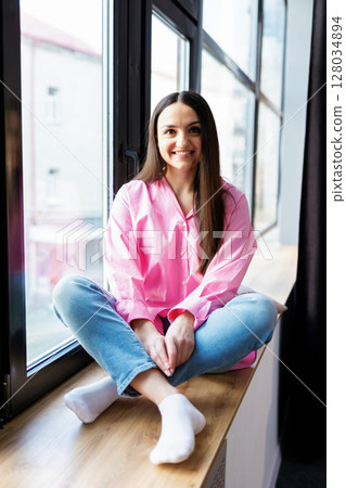 Pretty young woman sitting on windowsill relaxing at home looking at camera. Smiling calm lady chilling in apartment, dreaming, thinking of peaceful time enjoying peace of mind. 128034894