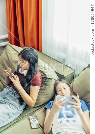 two girls relaxing on sofa using smartphone, headphones, casual home setting bright curtains and natural light window modern lifestyle, technology education entertainment, vertical two girls relaxing on sofa using smartphone, headphones, casual home setting bright curtains and natural light window modern lifestyle, technology education entertainment, vertical 128034947