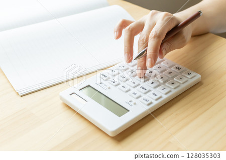 Calculator and notebook in the hands of a woman doing calculations 128035303