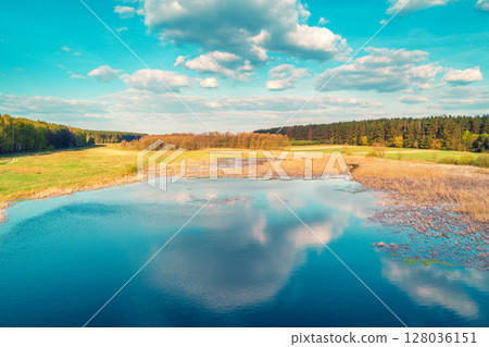 Aerial view of the countryside on a sunny day. Pond in a meadow surrounded by forest. Beautiful natural landscape with cloudy sky 128036151