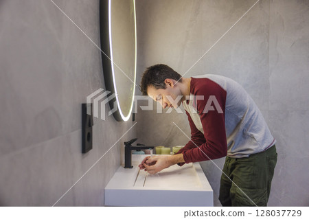 Young man washing his hands under running water in a modern bathroom, focusing on hygiene and cleanliness as part of a healthy daily routine. 128037729