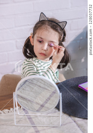 Little girl carefully applies pink eyeshadow in front of a mirror, wearing cat ears and focused on her makeup experiment Little girl carefully applies pink eyeshadow in front of a mirror, wearing cat ears and focused on her makeup experiment 128037732