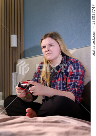 Teen girl sits barefoot on a bed with a wireless controller, showing a serious and competitive expression while playing a video game at home. 128037747