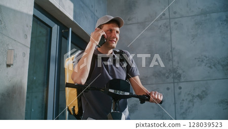 Delivery Man on Electric Scooter Standing near Office, Accepting New Order from Client Talking by Phone 128039523