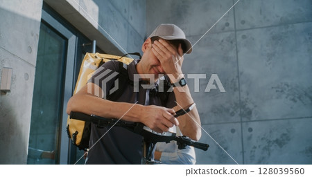 Tired Delivery Man Standing near Office, Searching for New Online Orders in App Using Smartphone Tired Delivery Man Standing near Office, Searching for New Online Orders in App Using Smartphone 128039560