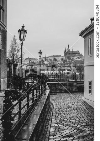 This black and white view captures Prague Castle rising majestically in the background, framed by lamp posts and a quiet cobblestone street lined with historic buildings. This black and white view captures Prague Castle rising majestically in the background, framed by lamp posts and a quiet cobblestone street lined with historic buildings. 128040804