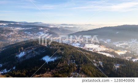 Stunning aerial view of Tanvald surrounded by snow-covered peaks in the Jizera Mountains. The serene winter landscape showcases valleys and dense forests under a clear sky. 128040806