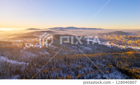 A peaceful winter evening unfolds over Jablonec nad Nisou with the snow-covered hills of the Jested Ridge in the background. The serene landscape captures the beauty of Czechia in winter. 128040807